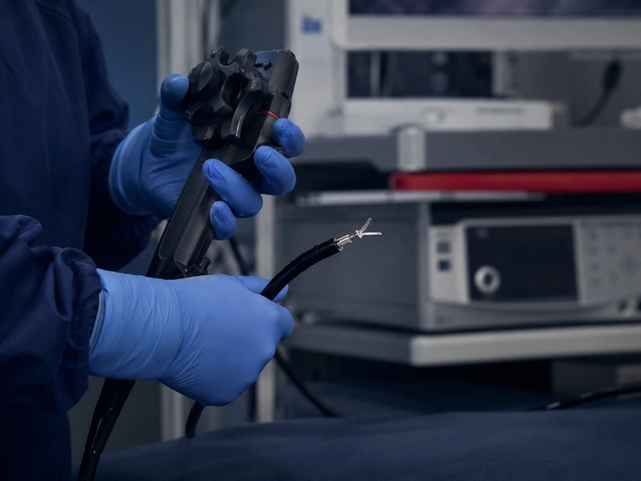 Mãos enluvadas no comando de um endoscópio, com o aplicador de clipe hemostático saindo da ponta distal e a torre de endoscopia desfocada ao fundo